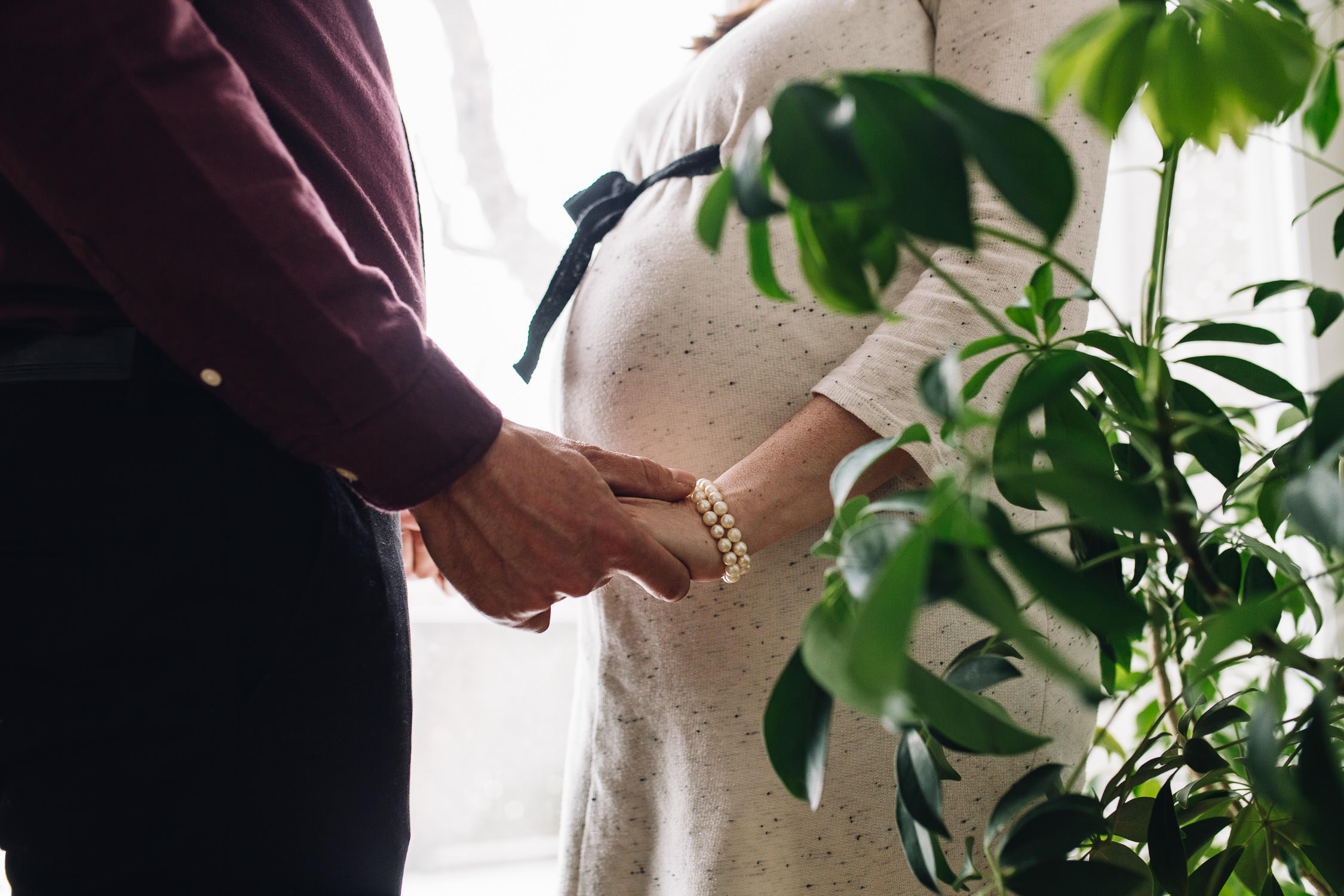 Un homme portant un haut bordeaux et une femme portant une robe blanche en tricot avec une cravate se tiennent par la main. Il y a une plante devant eux. L'image est prise de la poitrine vers le bas.