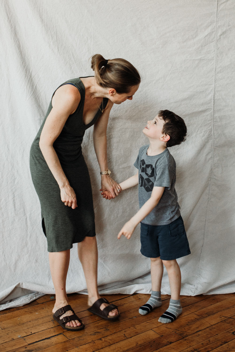 Woman wearing a green tank dress and birkenstock sandals is holding hands with a little boy wearing a grey t-shirt and shorts. He has brown hair and is wearing sneakers. They are standing in front of a beige backdrop and holding hands.