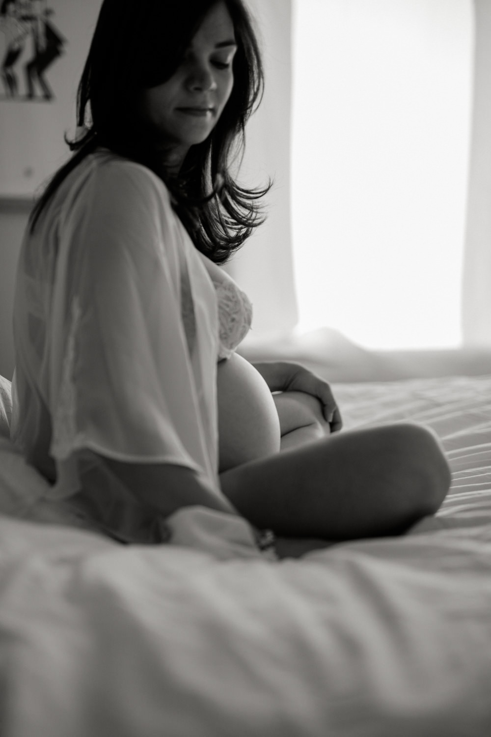 Pregnant woman wearing a lace bra and a robe is sitting crossed leg on the bed and looking down. You can see her pregnant belly.