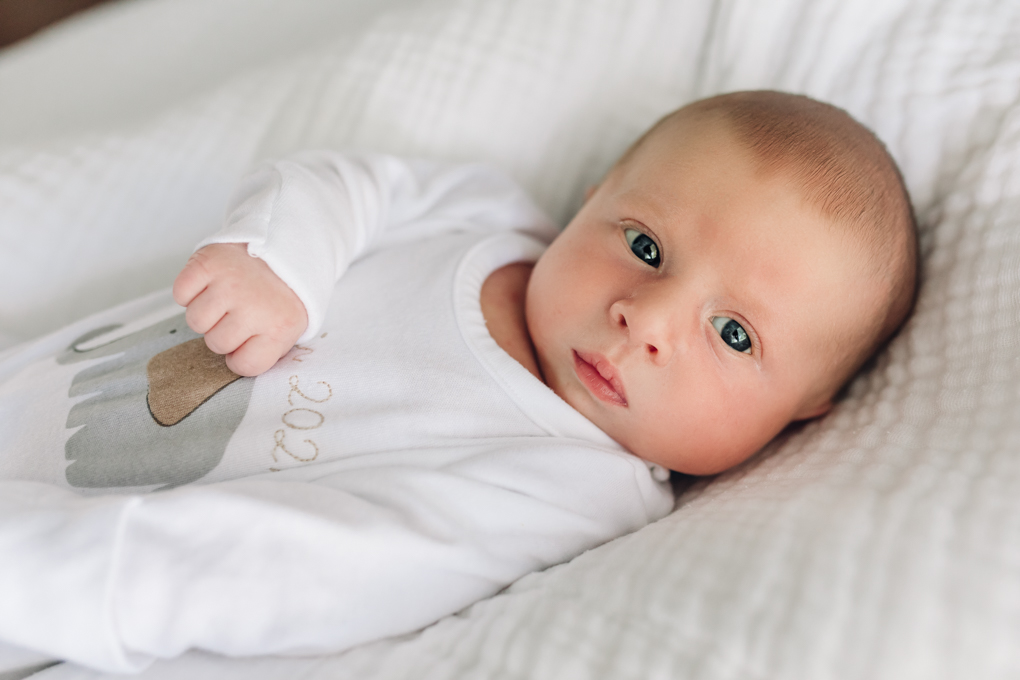 Un bébé vêtu d'une grenouillère blanche regarde par la fenêtre, les yeux grands ouverts.