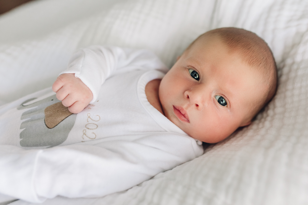 Baby in a white onesie is looking out the window with eyes wide open.