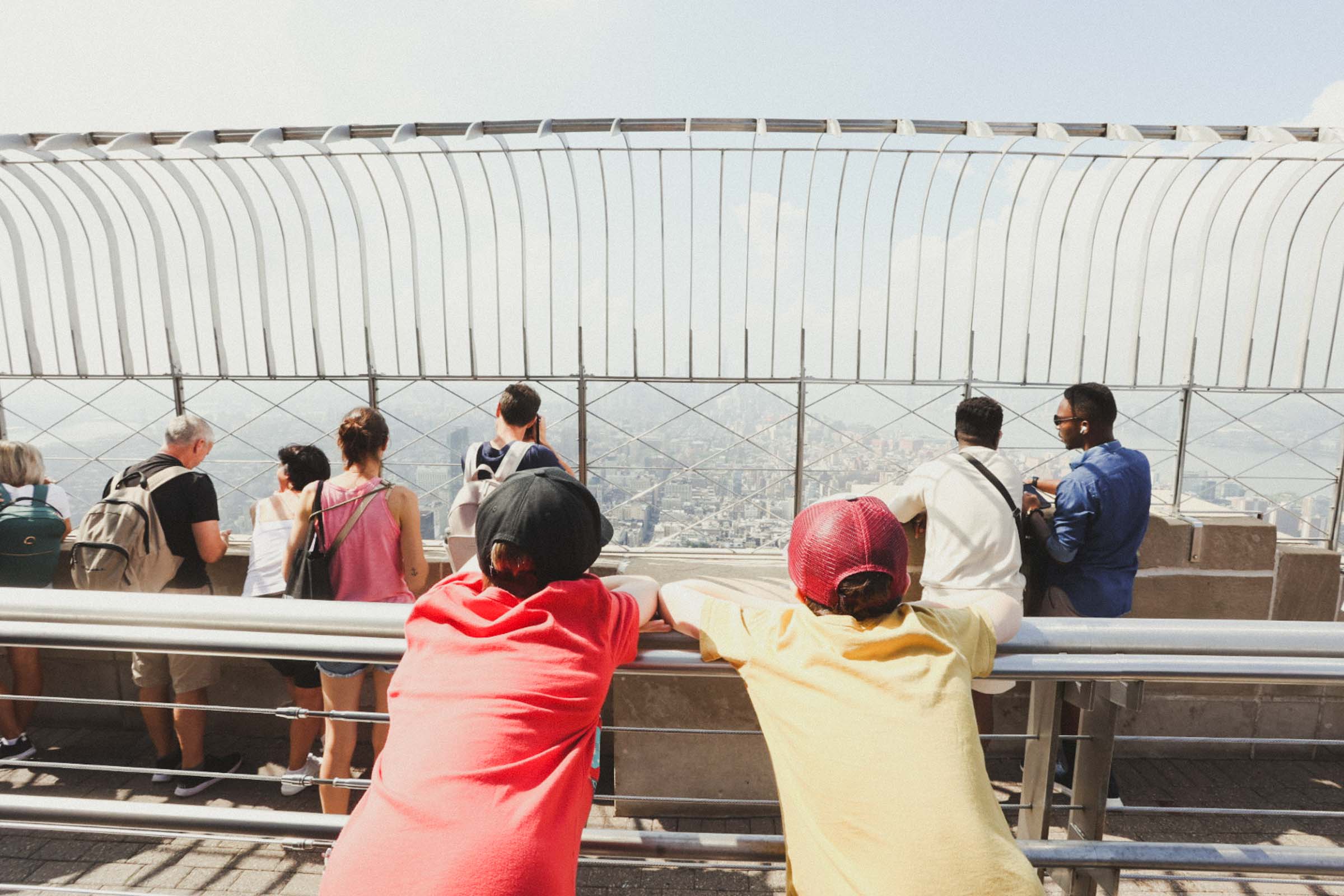 One boy wearing a red shirt and another boy wearing a yellow shirt leaning over a railing in the Empire State Building looking over Manhattan, NYC.