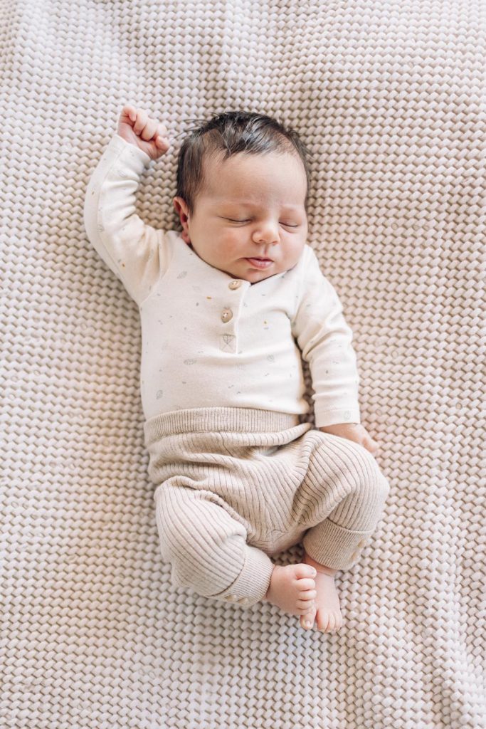 Newborn baby wearing long sleeve top and pants in an oatmeal colour is on an oatmeal coloured waffle blanket. He is asleep and one arm is up.