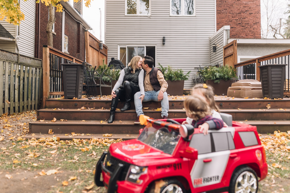 Maman et papa s'embrassent sous le porche arrière pendant que les enfants passent dans un camion de pompiers motorisé. Le petit garçon regarde les parents s'embrasser.