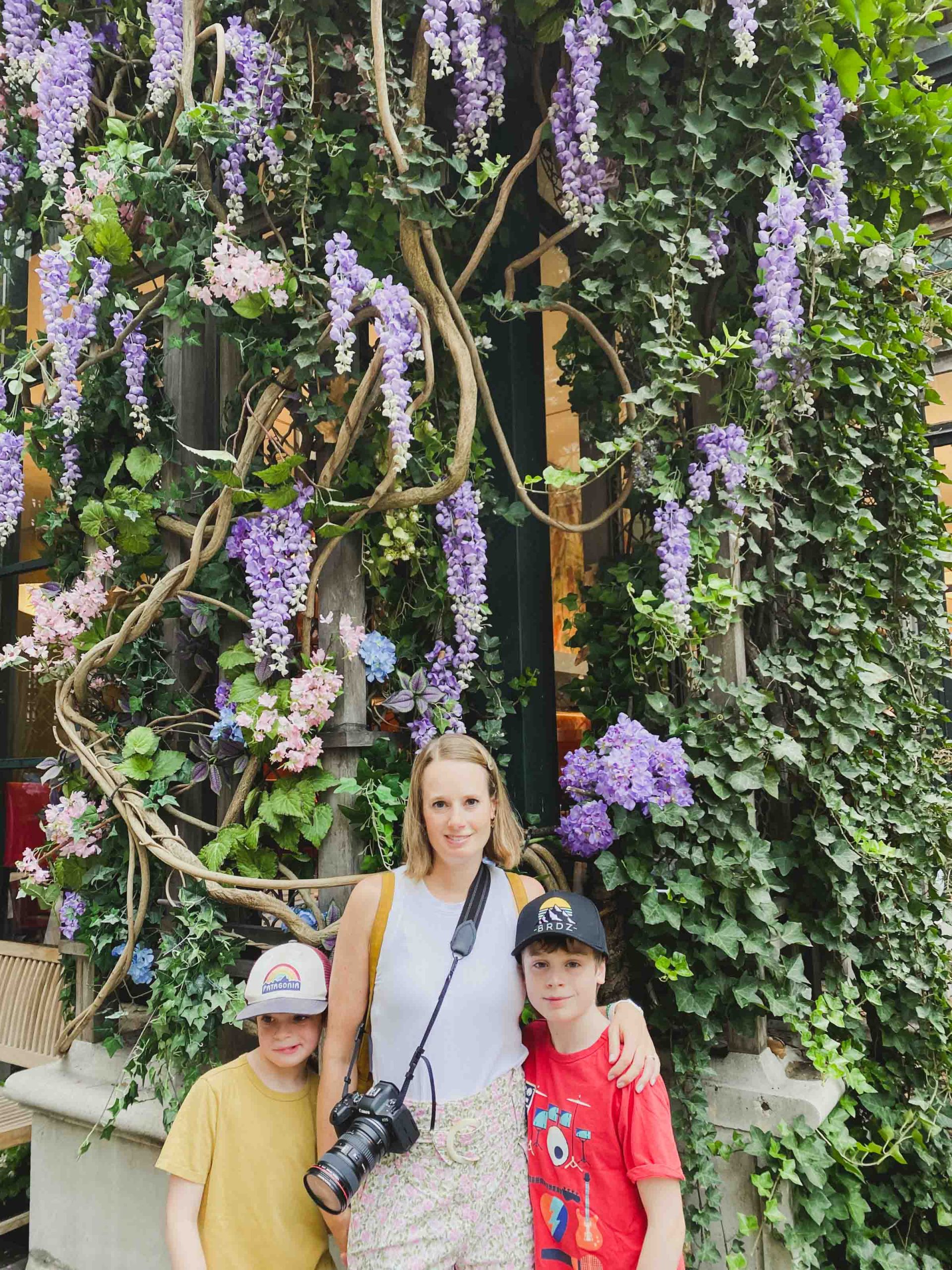 A mom with a huge camera and two boys age 10 and 8 standing close together in front of a wall of wisteria flowers.