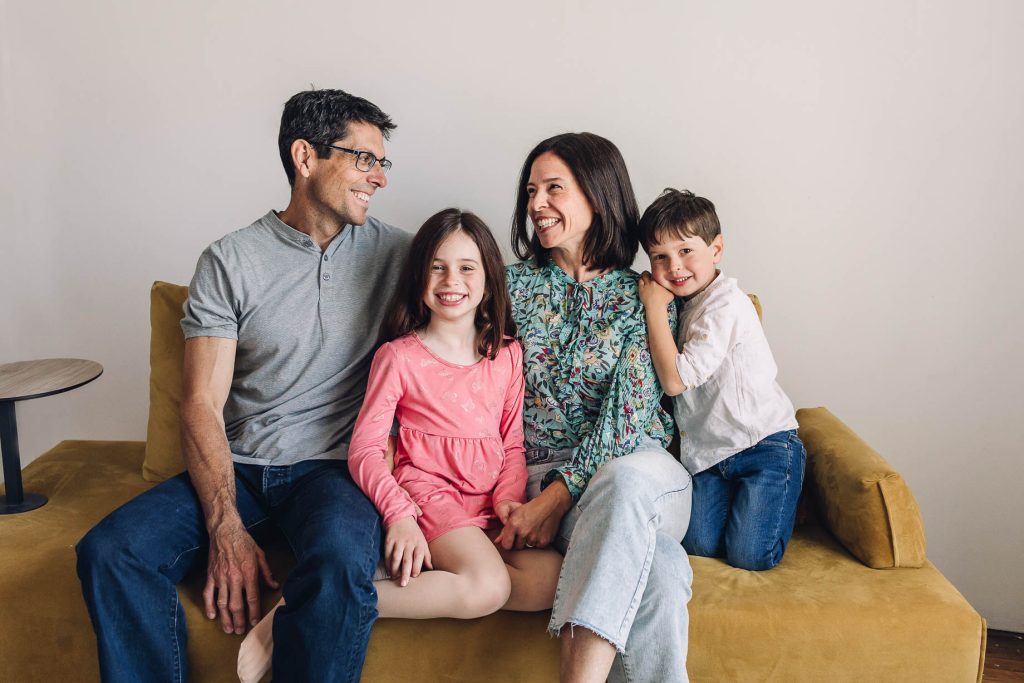 Family sitting on gold couch with mom and dad looking at each other smiling and kids smiling at camera by a Montreal Photographer.