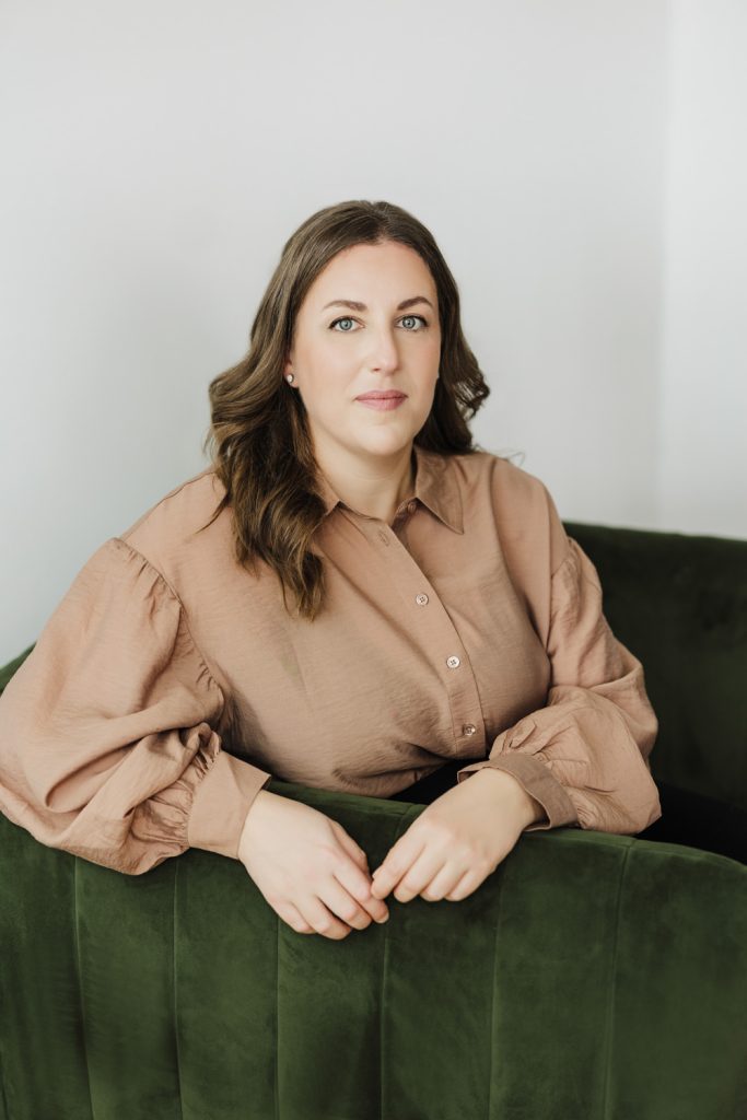 Lady wearing a brown top is leaning over a green chair and looking at the camera.