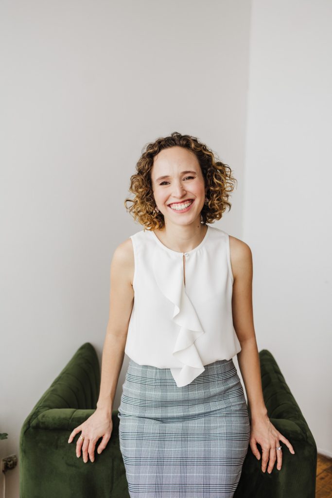 Lady with a tartan skirt and sleeveless white top is leaning against a green chair and smiling.