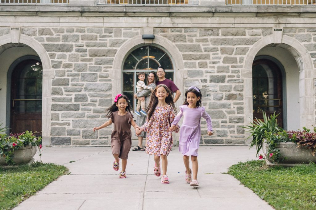 3 little girls holding hands and running towards the camera while their parents smile at them and mom holds their little brother