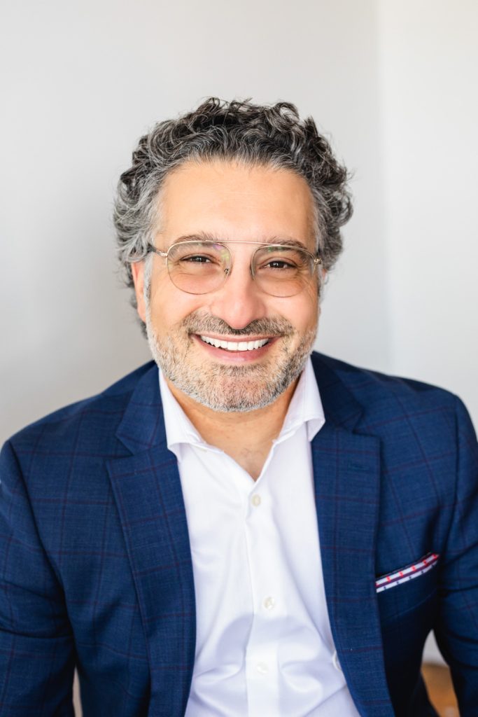 Man in a blue suite and white button up shirt is wearing glasses and has curly grey hair. He is looking at the camera.