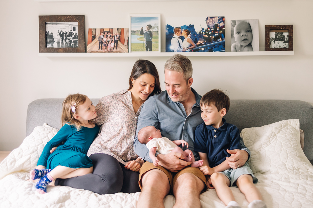 Family of two older children and parents sitting on bed and looking at a newborn baby.